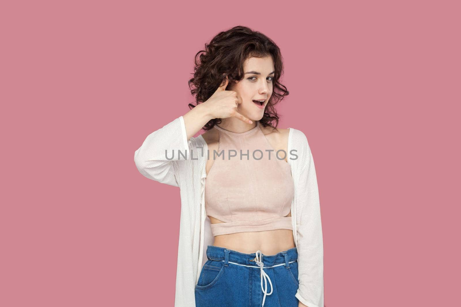 Portrait of funny beautiful brunette young woman with curly hairstyle in casual style standing with call hands gesture and looking at camera. indoor studio shot isolated on pink background.