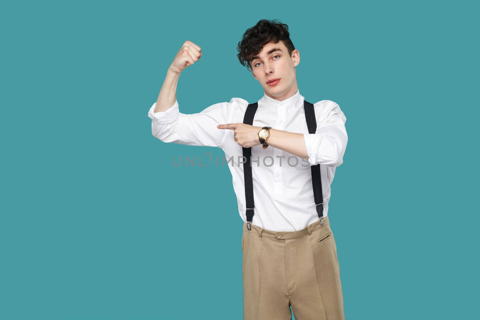 Proud man pointing at his biceps and looking at camera. Portrait of handsome hipster curly young businessman in classic casual white shirt and suspender. indoor studio shot isolated on blue background
