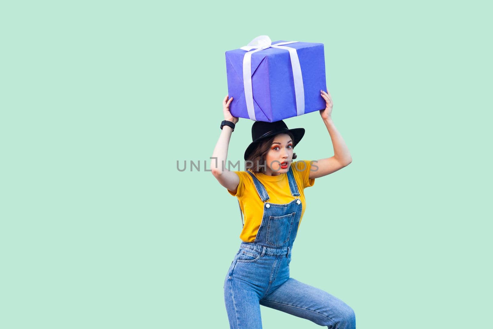 Suprised funny young girl in hipster wear in denim overalls and black hat standing and holding under head giant big heavy gift box with unbelievable face. Studio shot, green background, isolated