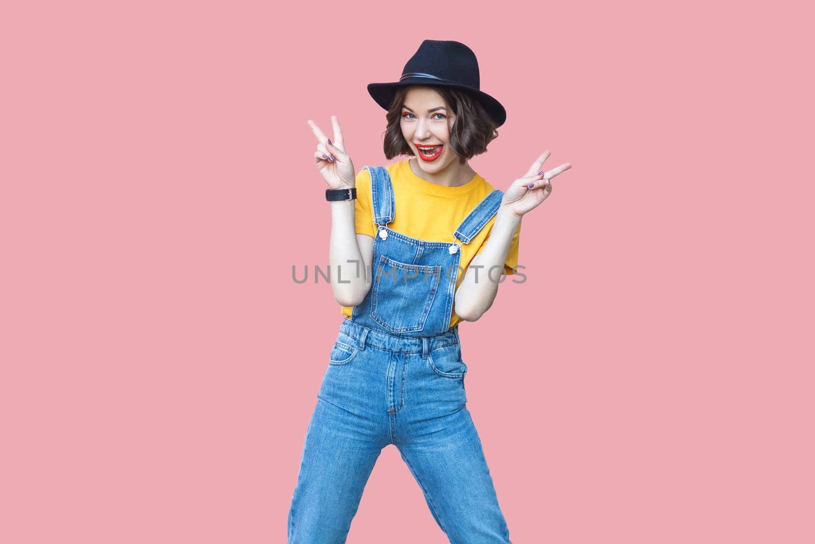 Portrait of excited beautiful young woman in yellow t-shirt and blue denim overalls with makeup and black hat standing and looking with victory or peace sign. studio shot isolated on pink background.