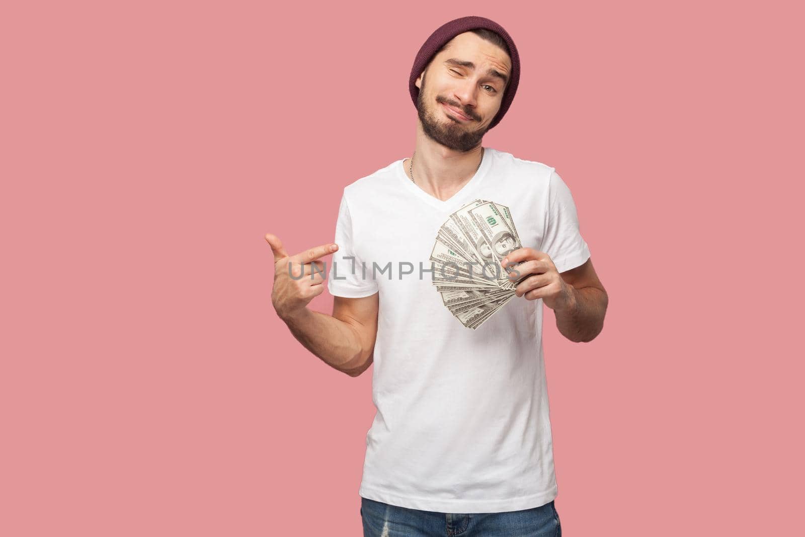I am wealthy man. Handsome bearded young hipster man in white shirt and casual hat, stand, holding fan of dollars money and pointing finger to cash. Indoor,isolated, studio shot, pink background