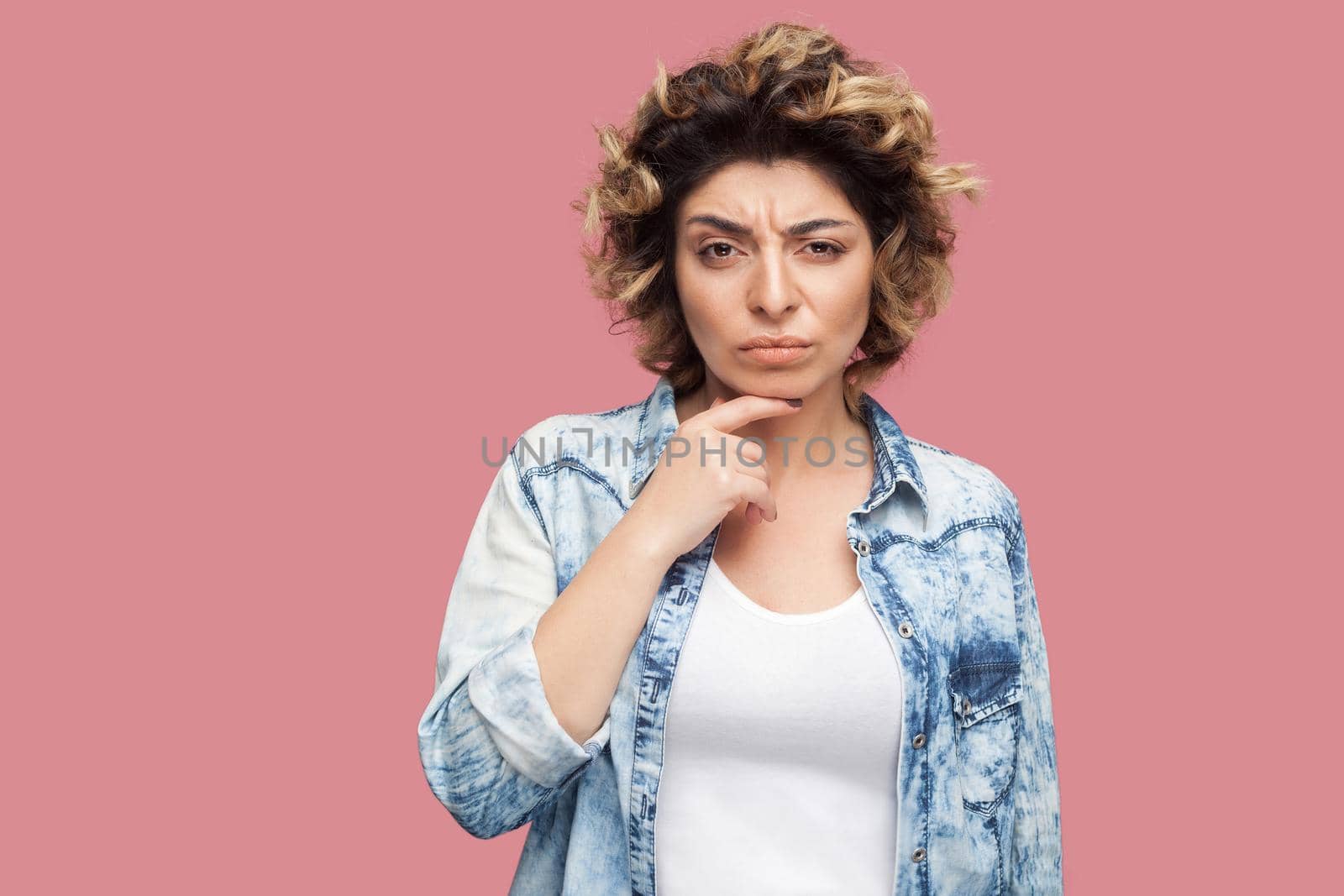Portrait of thoughtful young woman with curly hairstyle in casual style standing, touching her chin and looking at camera with confused thinking face. indoor studio shot, isolated on pink background.