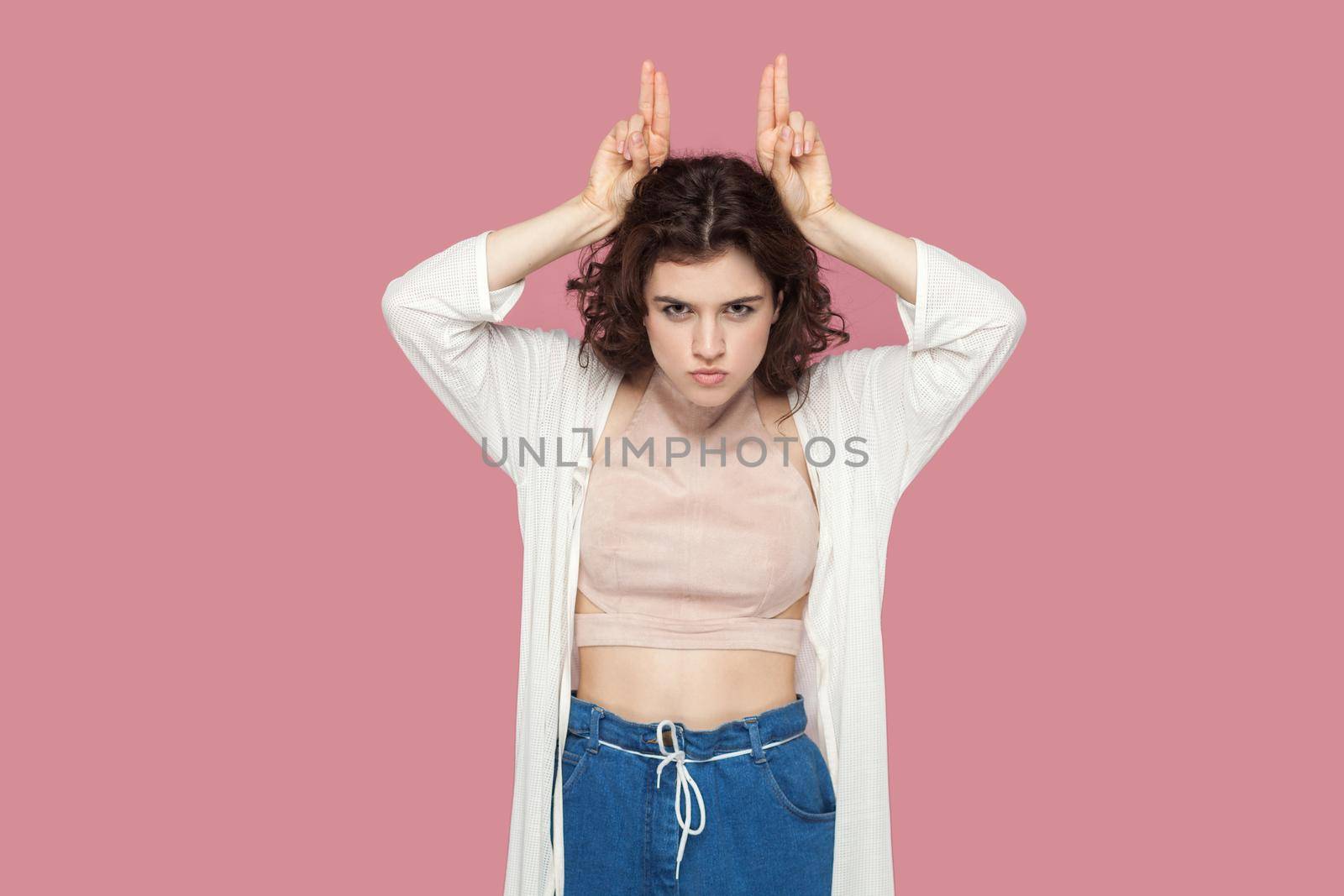 Portrait of serious funny cute brunette young woman with curly hairstyle in casual style standing with finger horns on her head and looking at camera. indoor studio shot isolated on pink background.