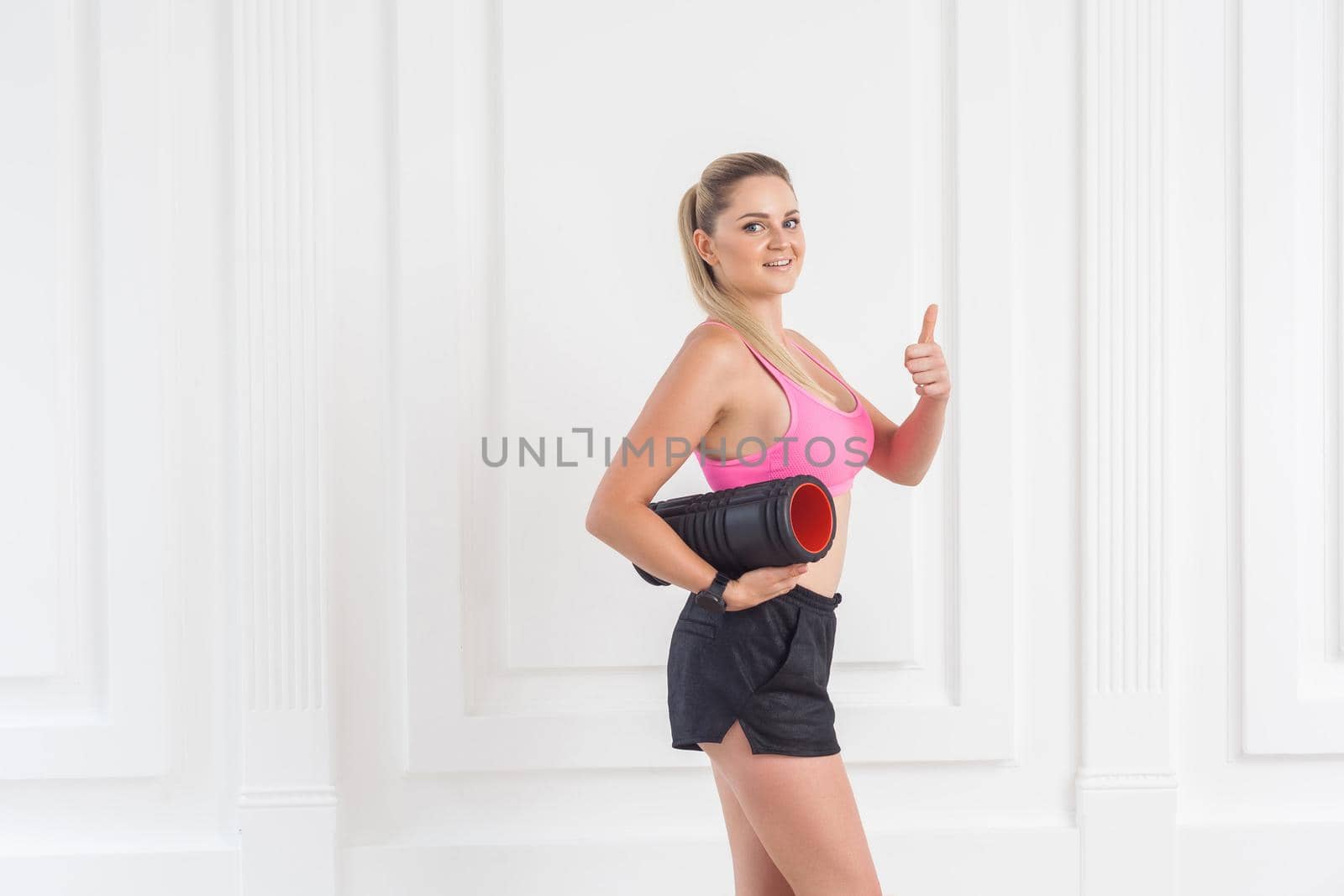 Sporty concept. Satisfied athletic young beautiful blonde girl in pink top and black shorts standing, holding foam roll and showing thumbs up. Studio shot, looking at camera, indoor, isolated