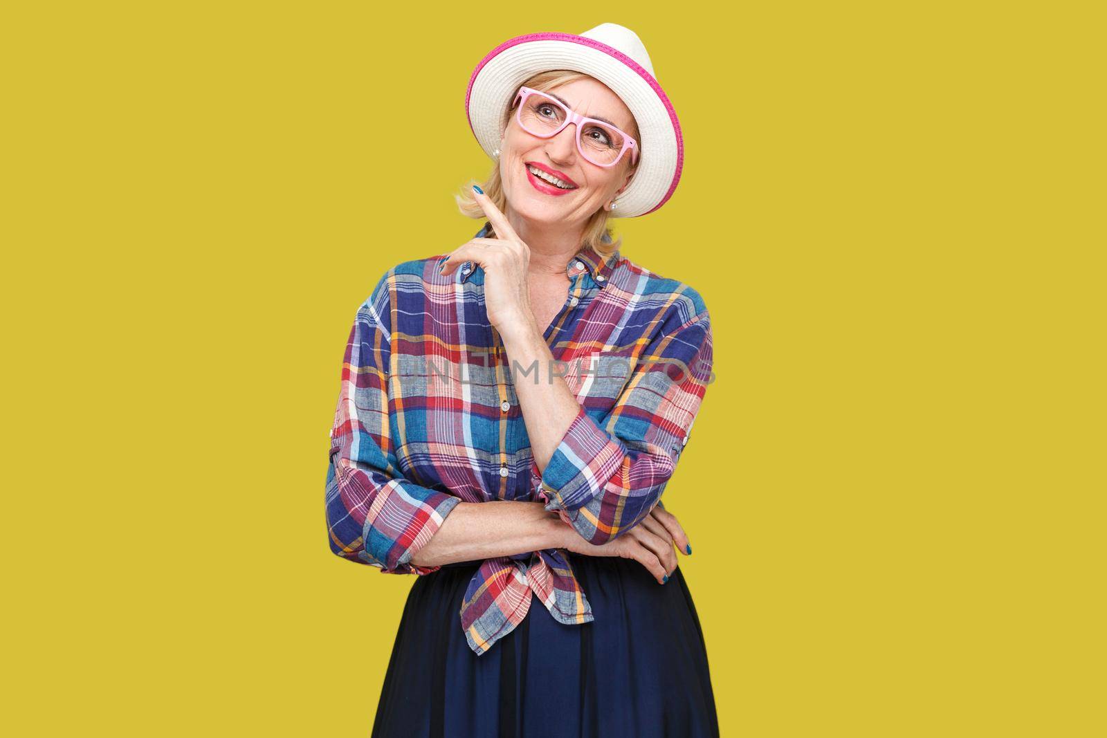 dreaming about something beautiful. Portrait of toothy smiley modern stylish mature woman in casual style with hat standing, looking away and dreaming. indoor studio shot isolated on yellow background