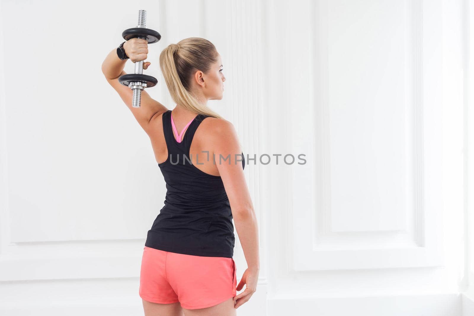 Striving for strong. Backside of young adult motivation athletic bodybuilder woman in pink shorts and black top standing, holding dumbells doing exercise for arms at the gym with perfect figure.