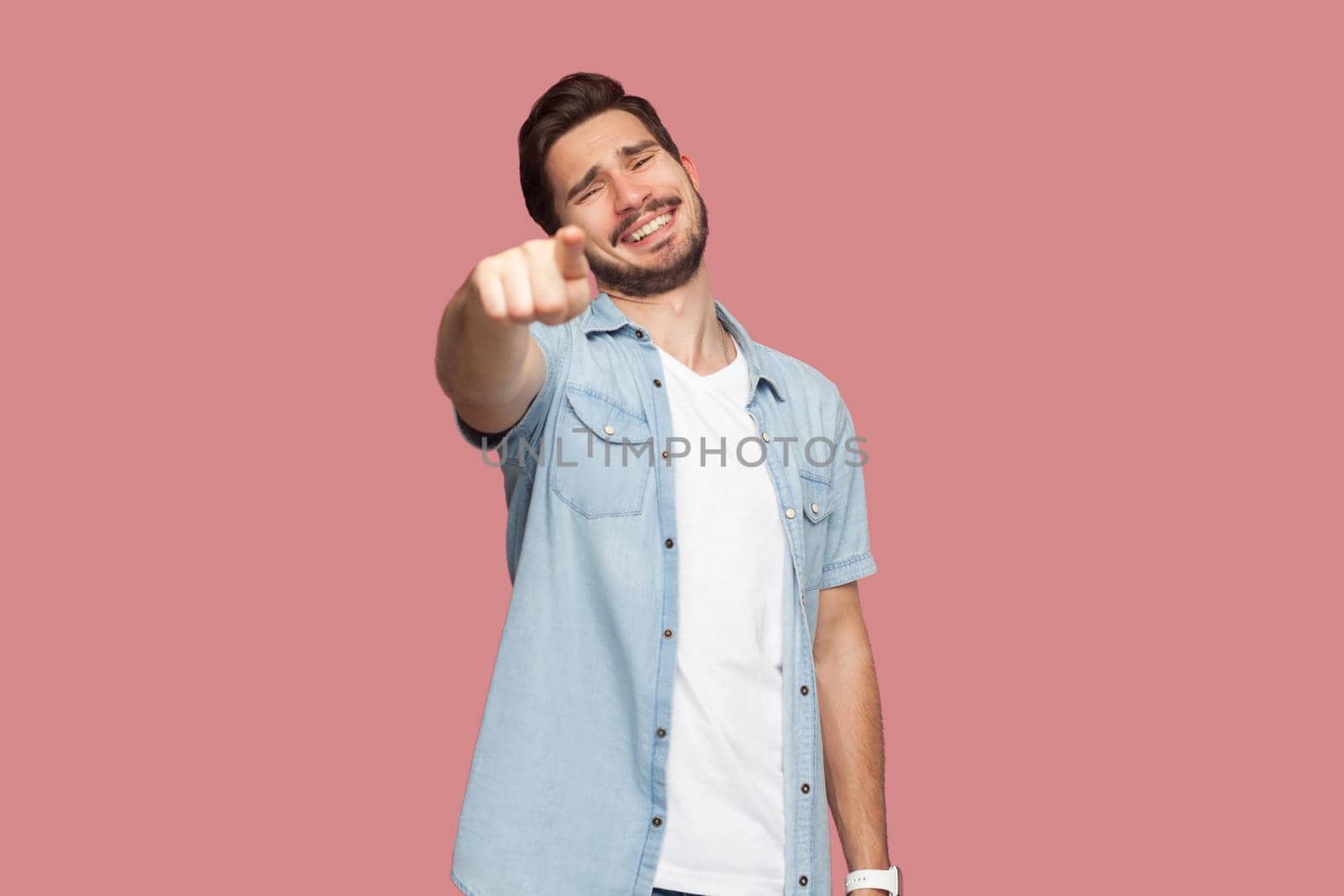 Portrait of funny happy handsome bearded young man in blue casual style shirt standing pointing and looking at camera with toothy smile. indoor studio shot, isolated on pink background.