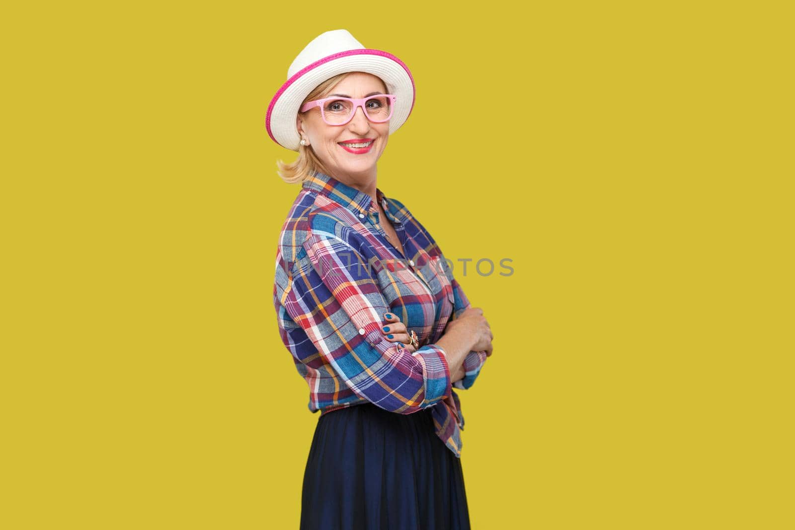 Portrait of happy modern stylish mature woman in casual style with hat and eyeglasses standing, crossed arms and looking at camera with toothy smile. indoor studio shot isolated on yellow background.