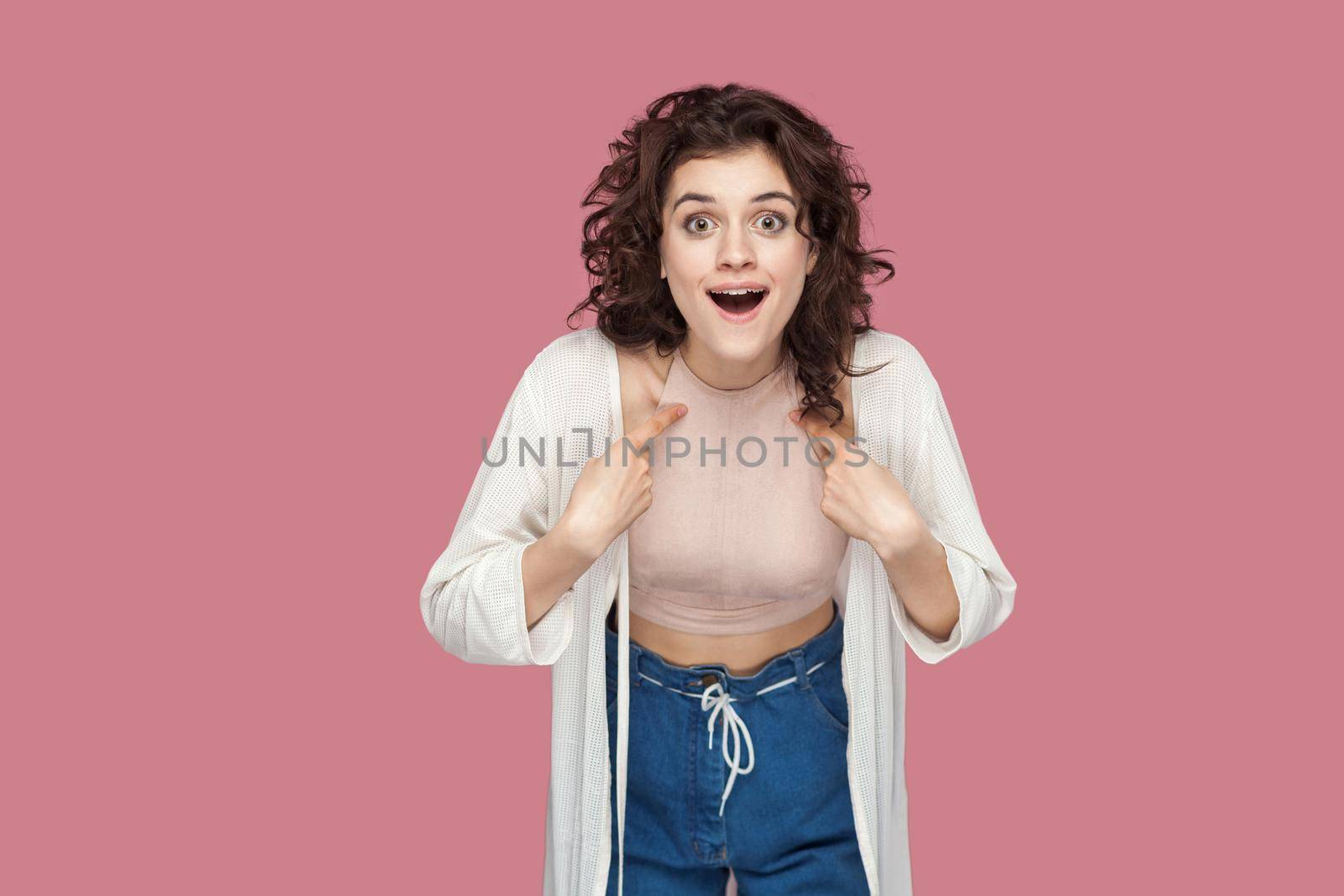 me? Portrait of surprised beautiful brunette young girl with curly hairstyle in casual style pointing herself, asking and looking at camera with amazed face. studio shot isolated on pink background.