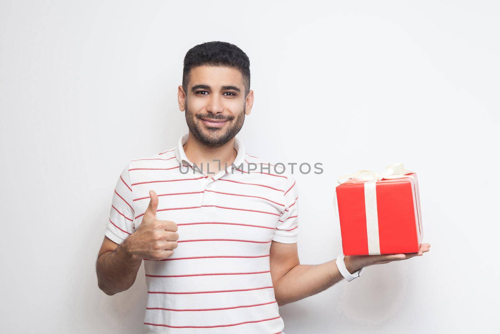 Like it! Satisfied handsome modern young adult man in t-shirt standing, holding big gift box and showing finger up with smile, looking at camera. Studio shot, white background, isolated, indoor