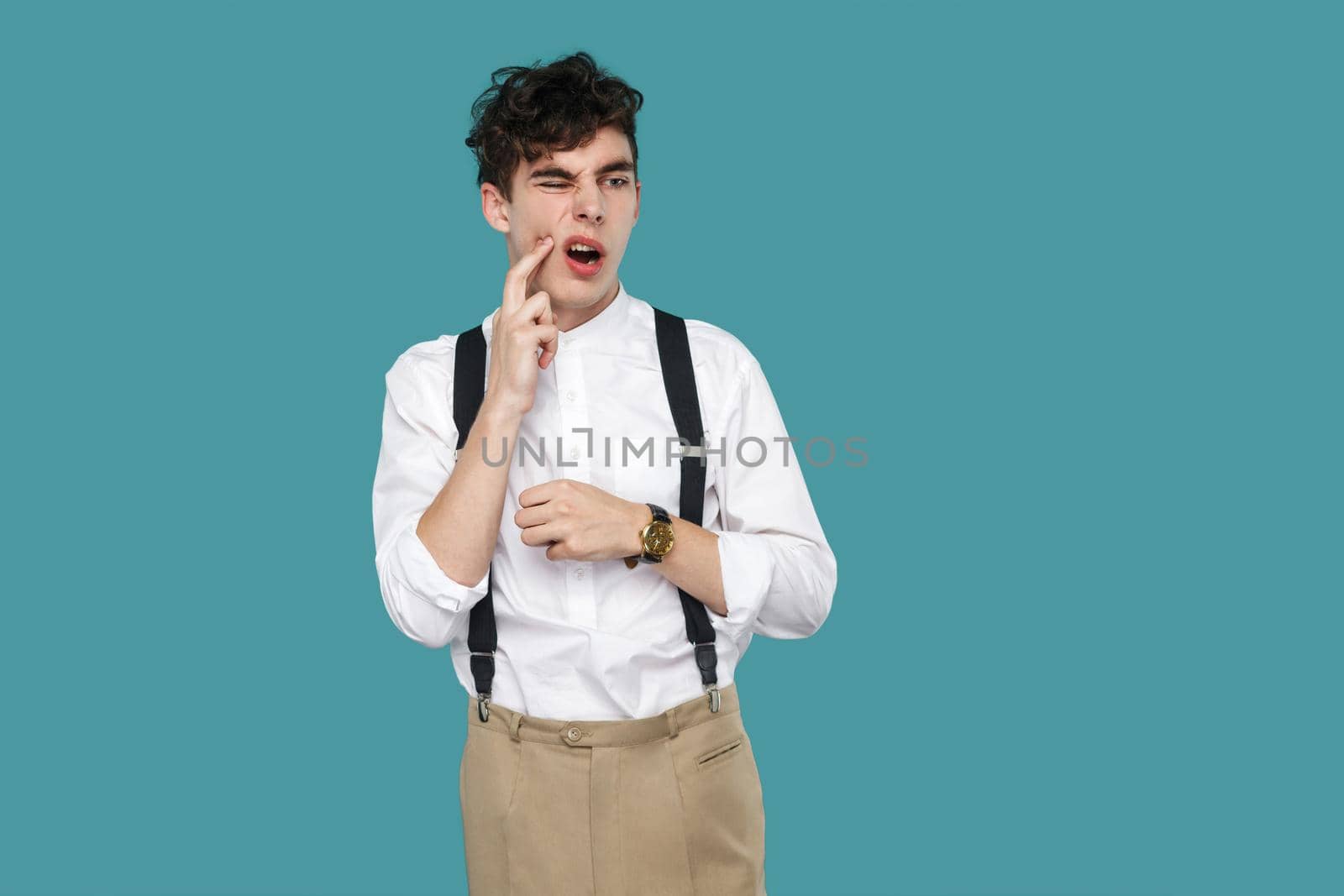 Tooth pain. man touching his chin because toothache. Portrait of handsome hipster curly young businessman in classic casual white shirt and suspender standing. studio shot isolated on blue background.