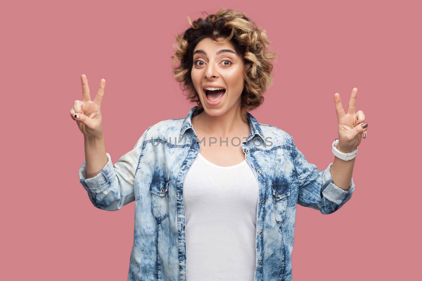 Portrait of happy funny young woman with curly hair in casual shirt standing with victory or peace sign gesture and looking at camera with amazed face. indoor studio shot, isolated on pink background.