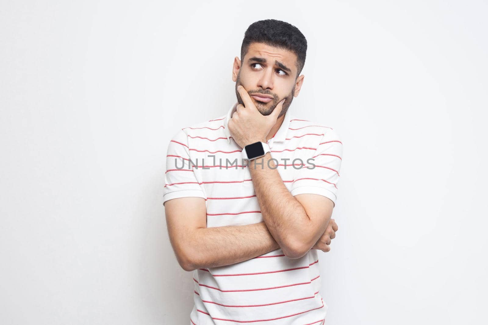 Portrait of pensive handsome bearded young man in striped t-shirt standing, touching his face, looking aside away and thinking about something. indoor studio shot, isolated on white background.