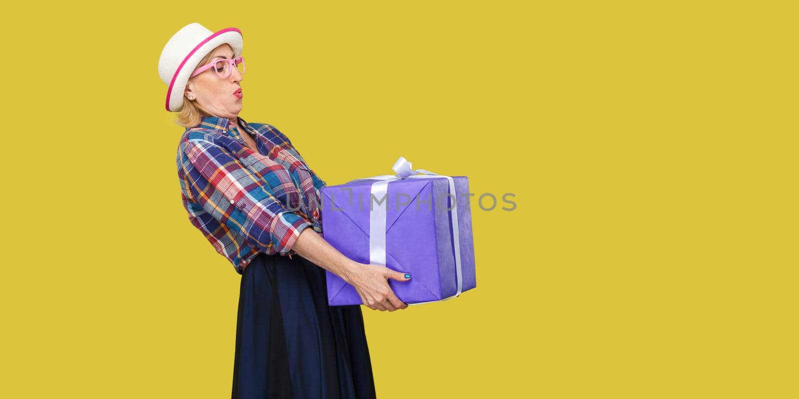 Side view portrait of cheerful wondering modern grandma in white hat and checkered shirt standing and trying to hold giant big heavy gift box and greeting you. Studio shot, yellow background, isolated