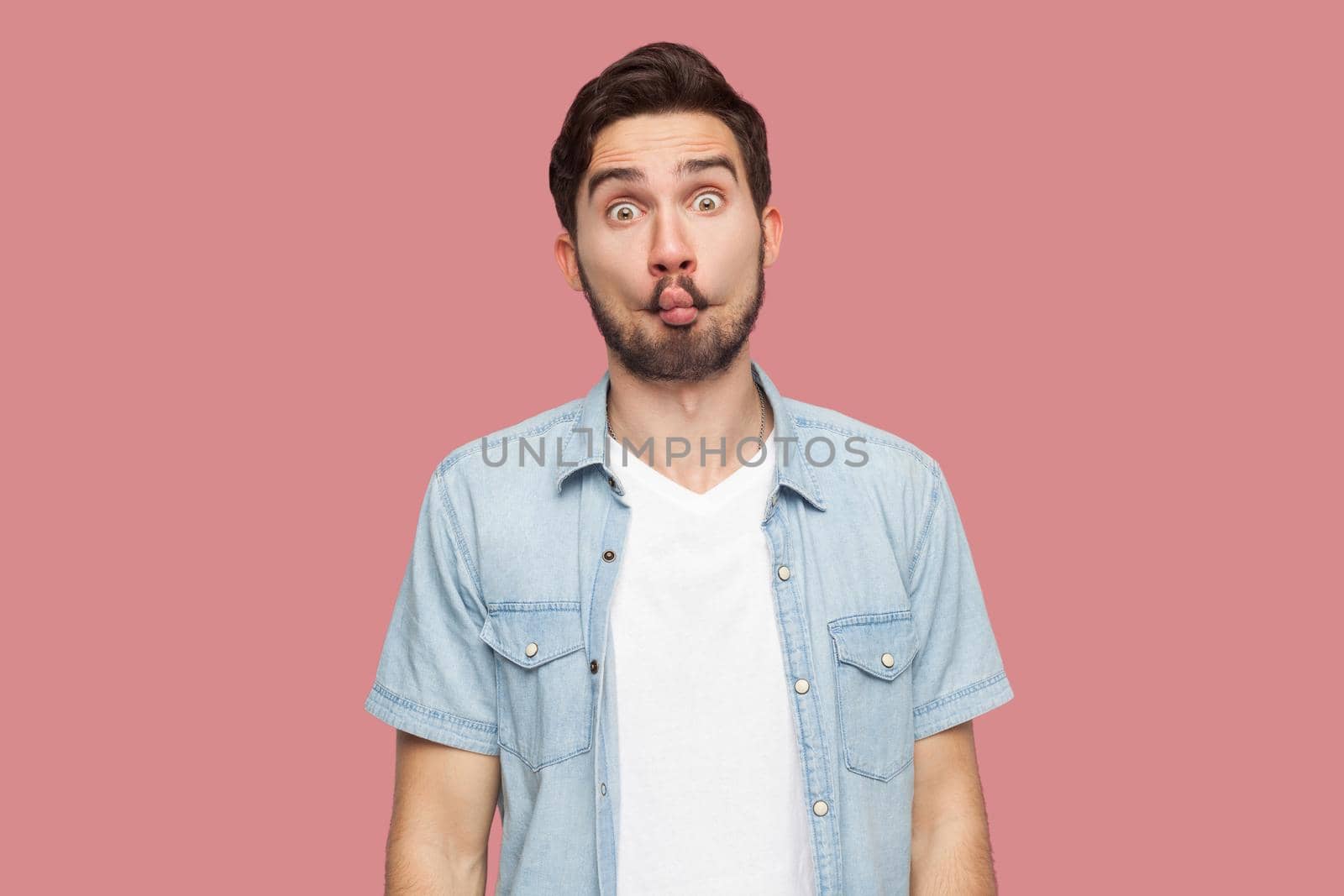Portrait of funny comic face handsome bearded young man in blue casual style shirt standing with fish lips and looking at camera. indoor studio shot, isolated on pink background.