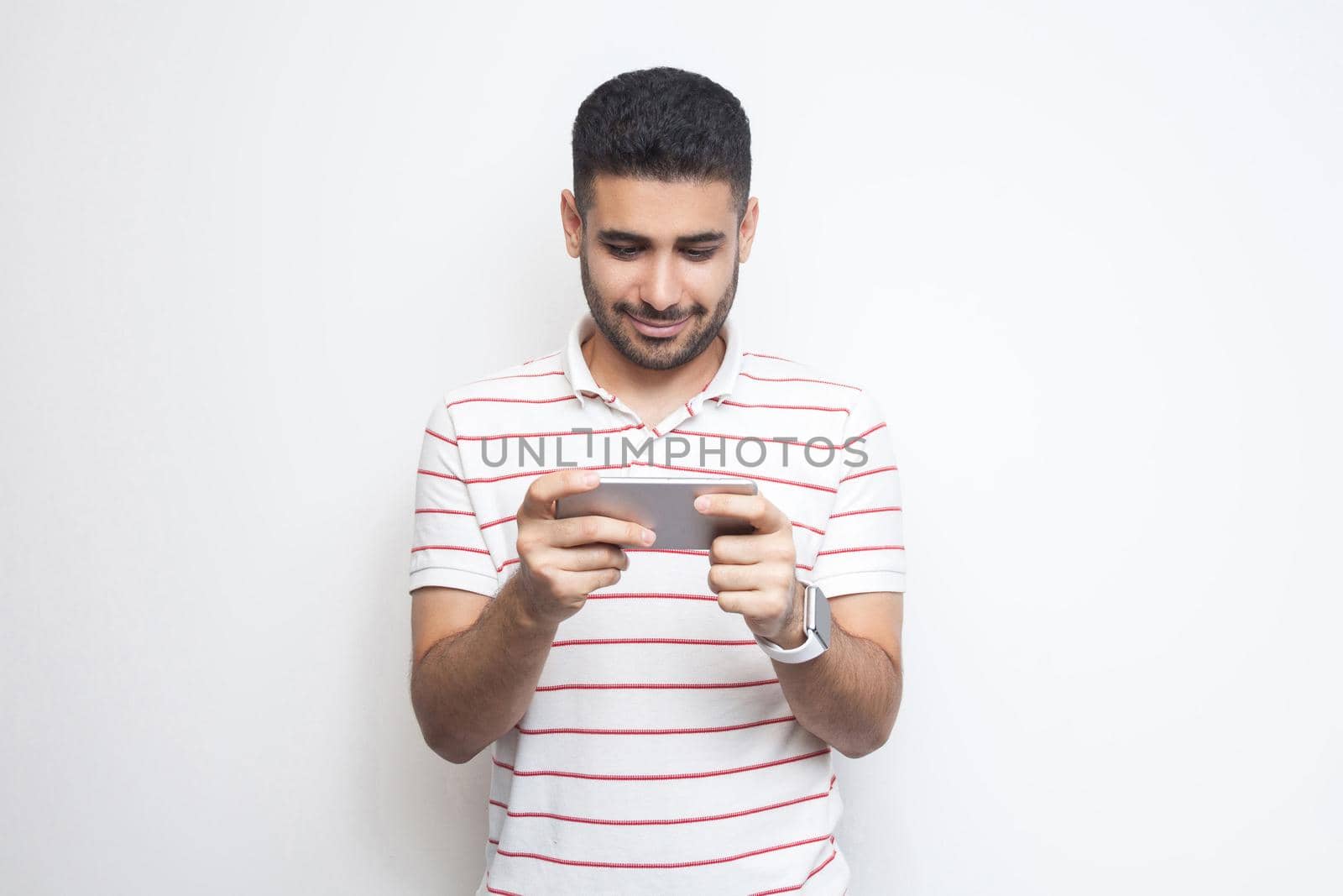 Portrait of happy handsome blogger young adult man wearing T-shirt standing, using smartphone and texting message with smile, looking video. Indoor, isolated, studio shot, white background