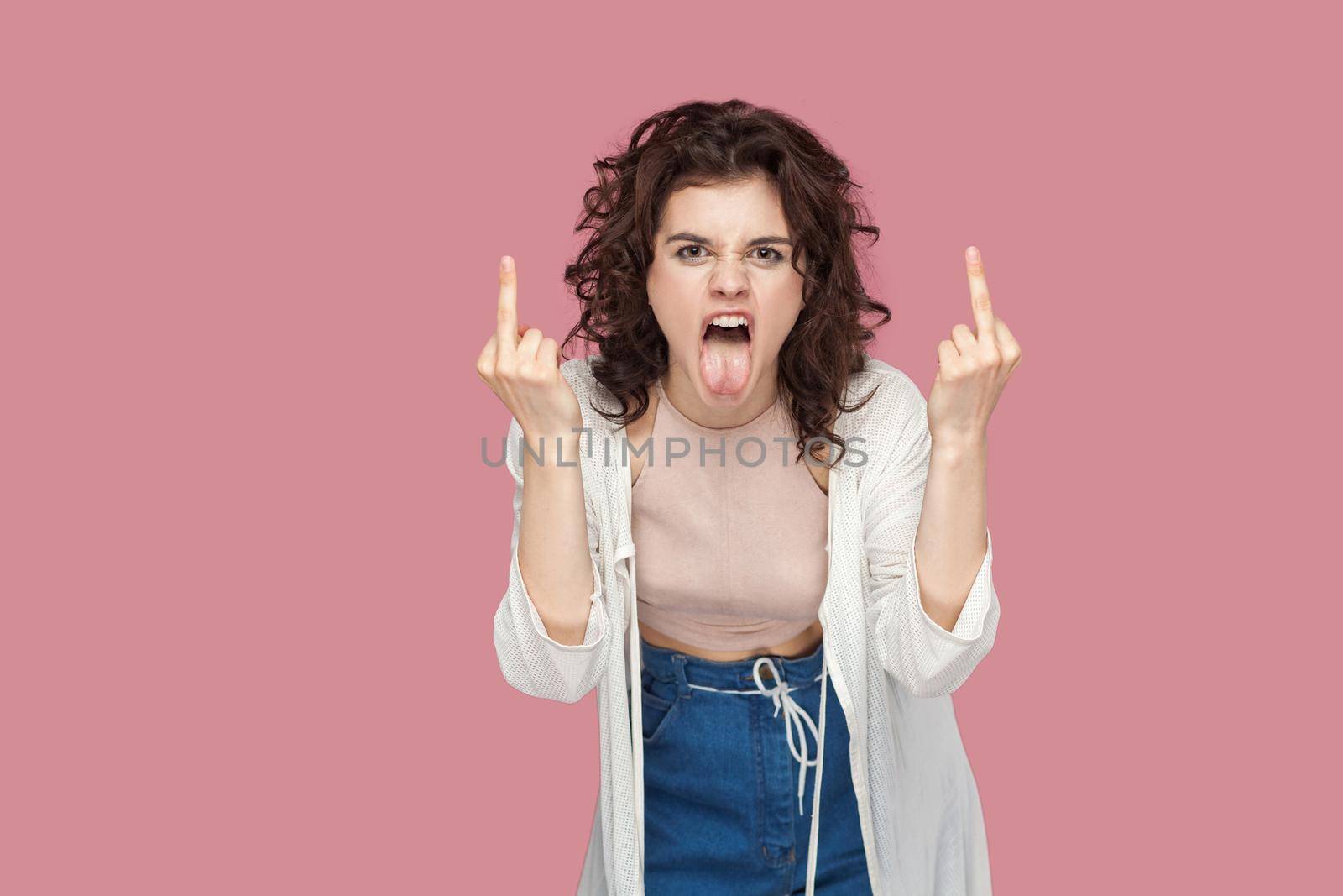 Portrait of angry screaming brunette young woman with curly hairstyle in casual style standing and looking at camera with middle finger fuck sign. indoor studio shot isolated on pink background.