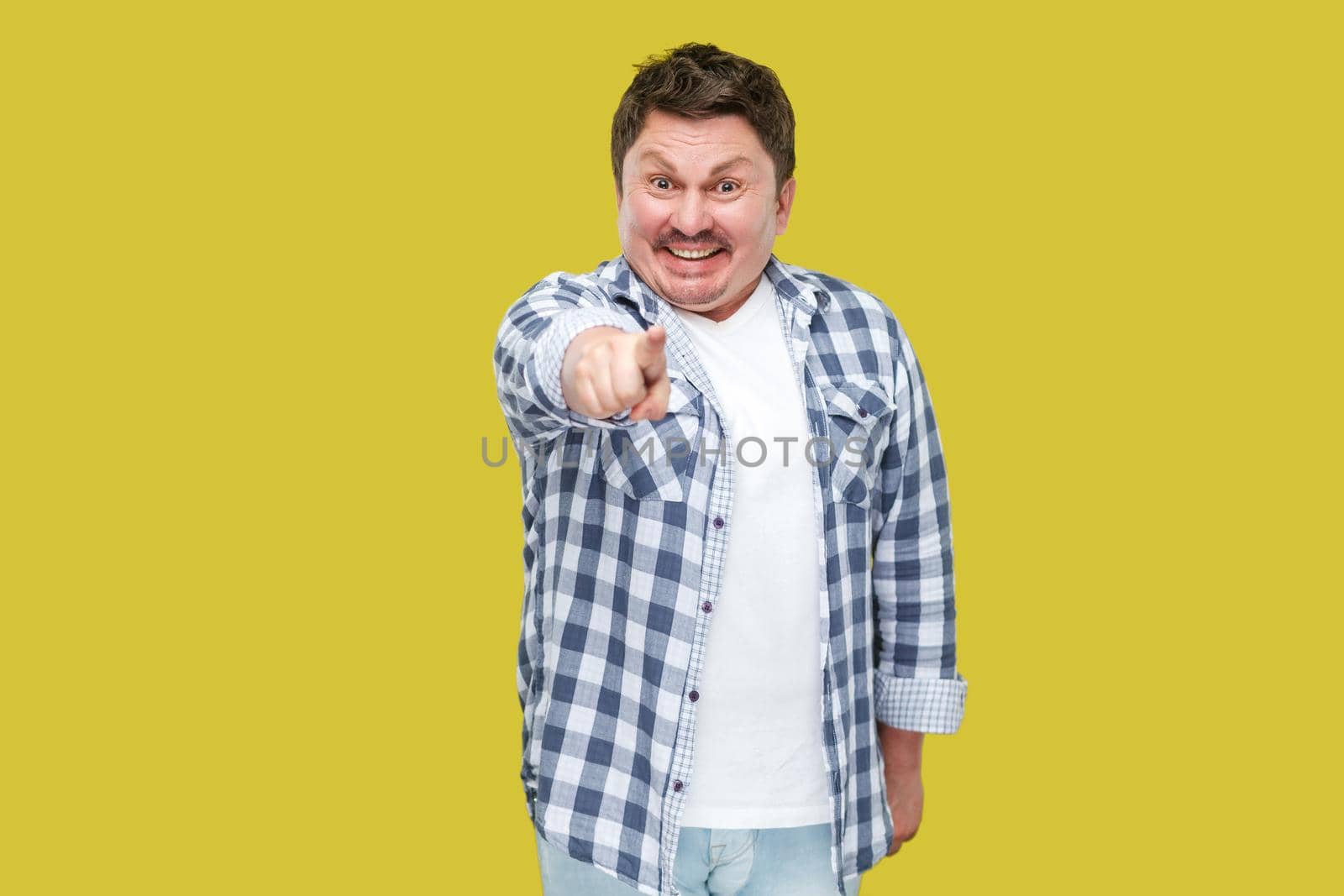 Portrait of excited happy handsome middle aged business man in casual checkered shirt standing, pointing and looking at camera with toothy smile. indoor studio shot, isolated on yellow background.