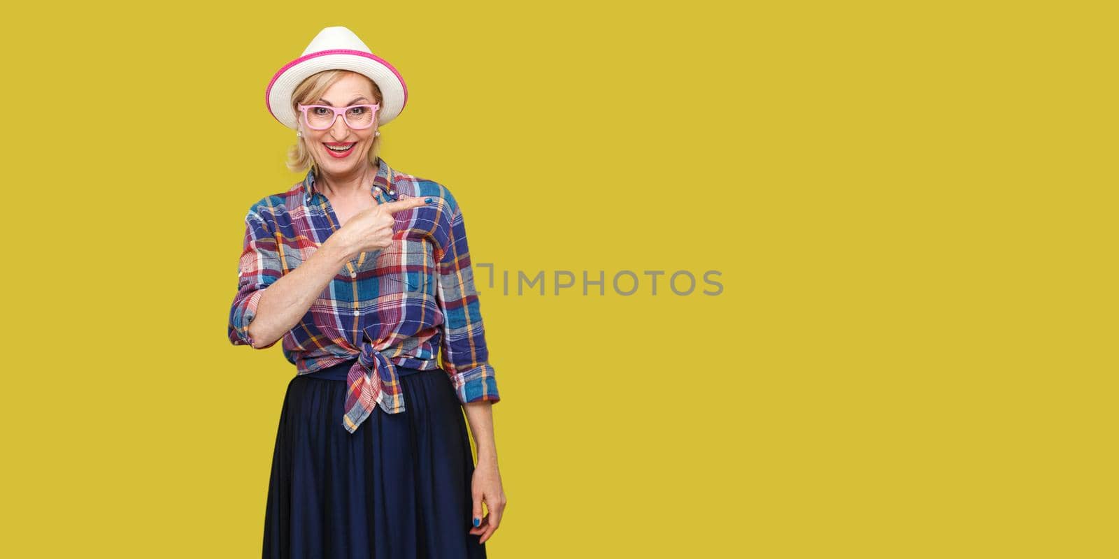 Portrait of happy modern stylish mature woman in casual style with hat and eyeglasses standing looking at camera and pointing and wall empty copyspace. indoor studio shot isolated on yellow background