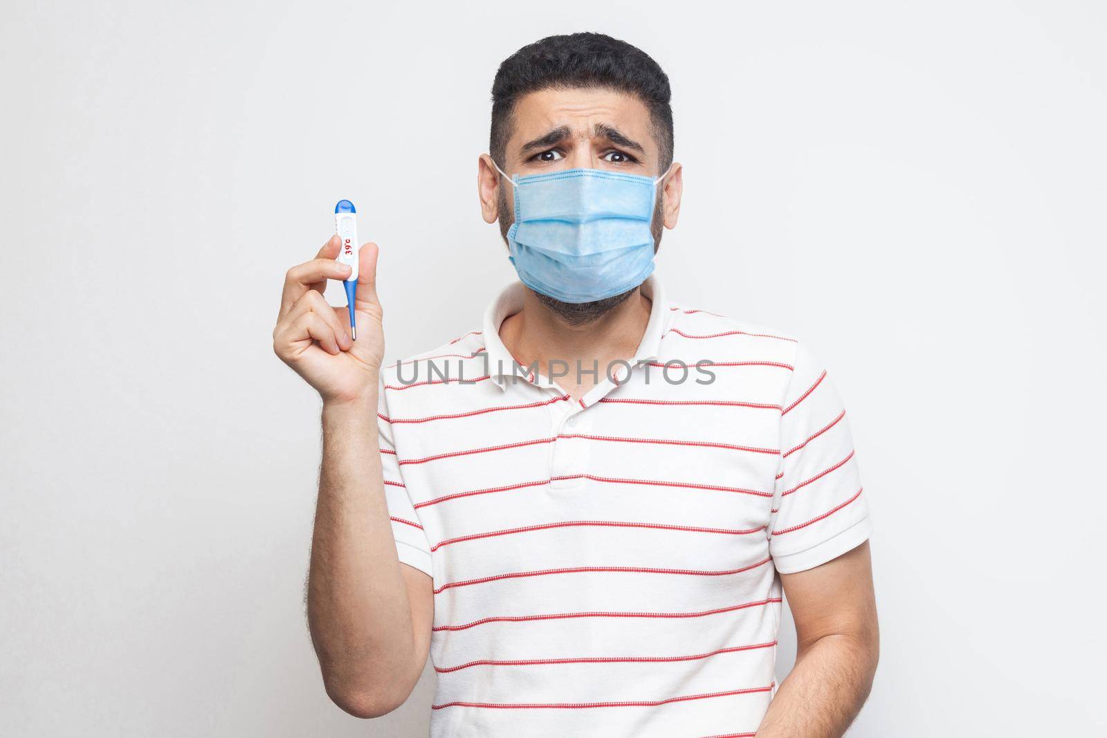 39 degress. Scared panic worry sick man with surgical medical mask standing, holding thermometer are shoched for high temperature, looking at camera. Indoor, isolated, white background, studio shot