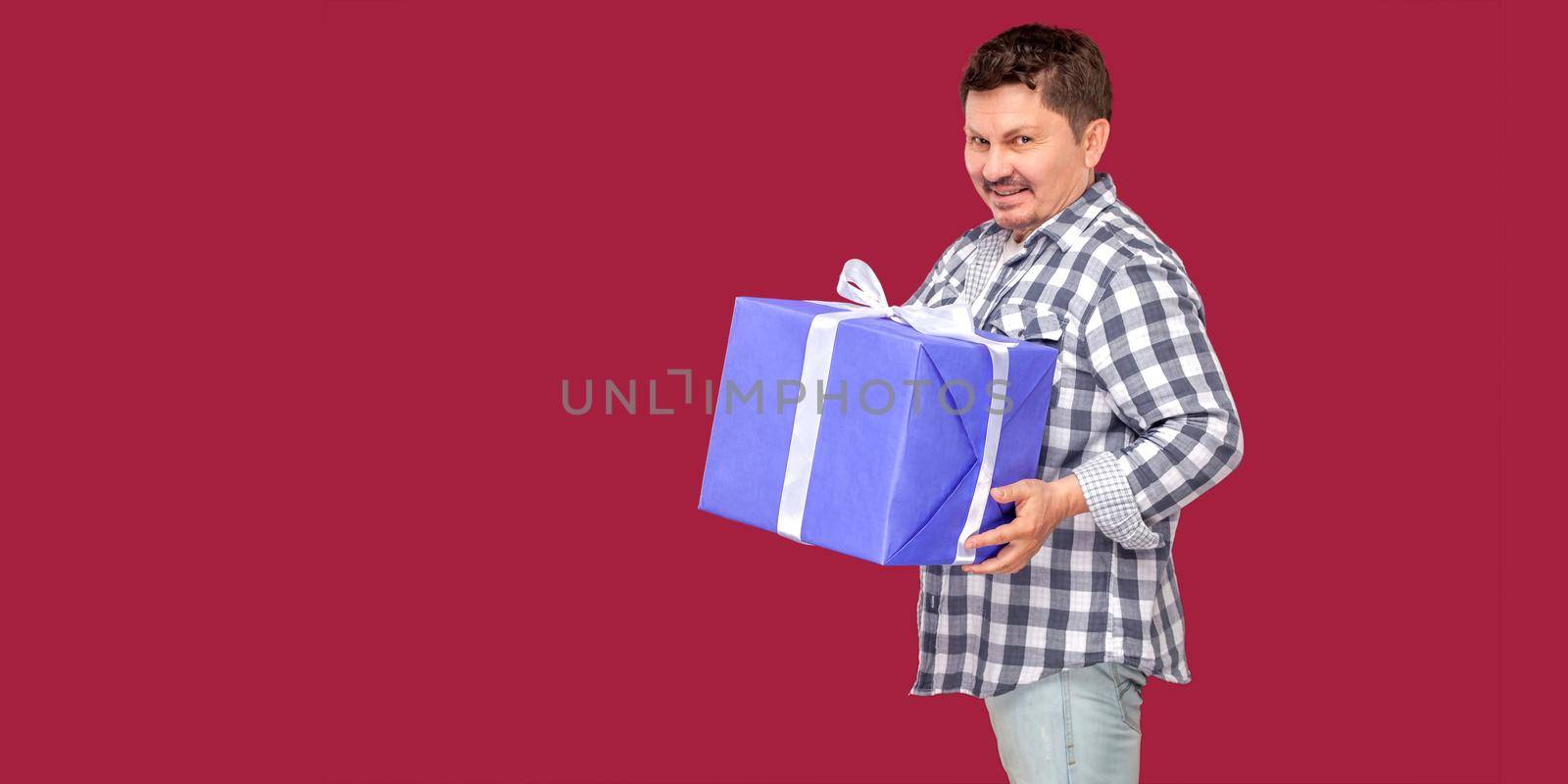 Portrait of happy modern middle aged man in white t-shirt and in checkered shirt standing and holding big heavy gift box with smile, looking at camera. indoor studio shot on isolated red background.
