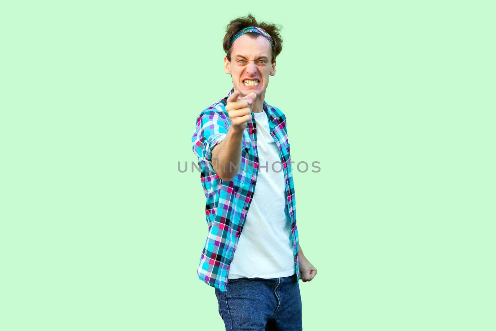 Portrait of angry nervous young man in casual blue checkered shirt and headband standing, clenching teeth, blaming and looking at camera. indoor studio shot, isolated on light green background.