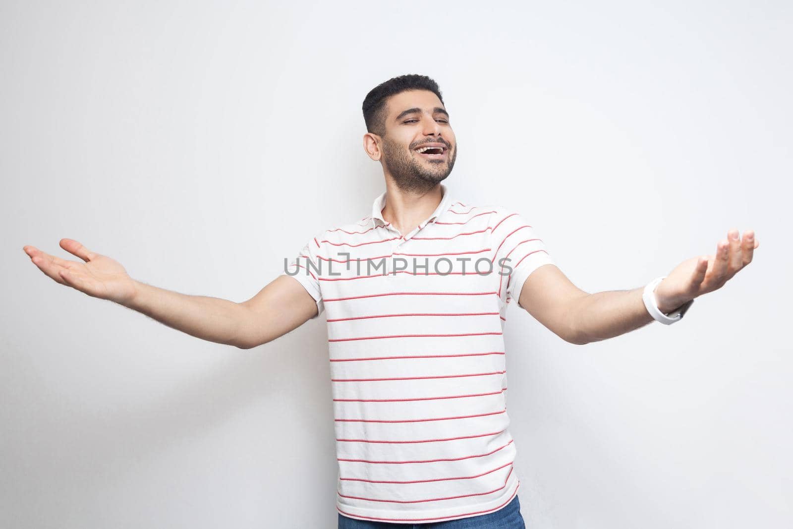 Portrait of happy handsome bearded young man in striped t-shirt standing with wide raised arms and looking at camera with toothy smile. indoor studio shot, isolated on white background.