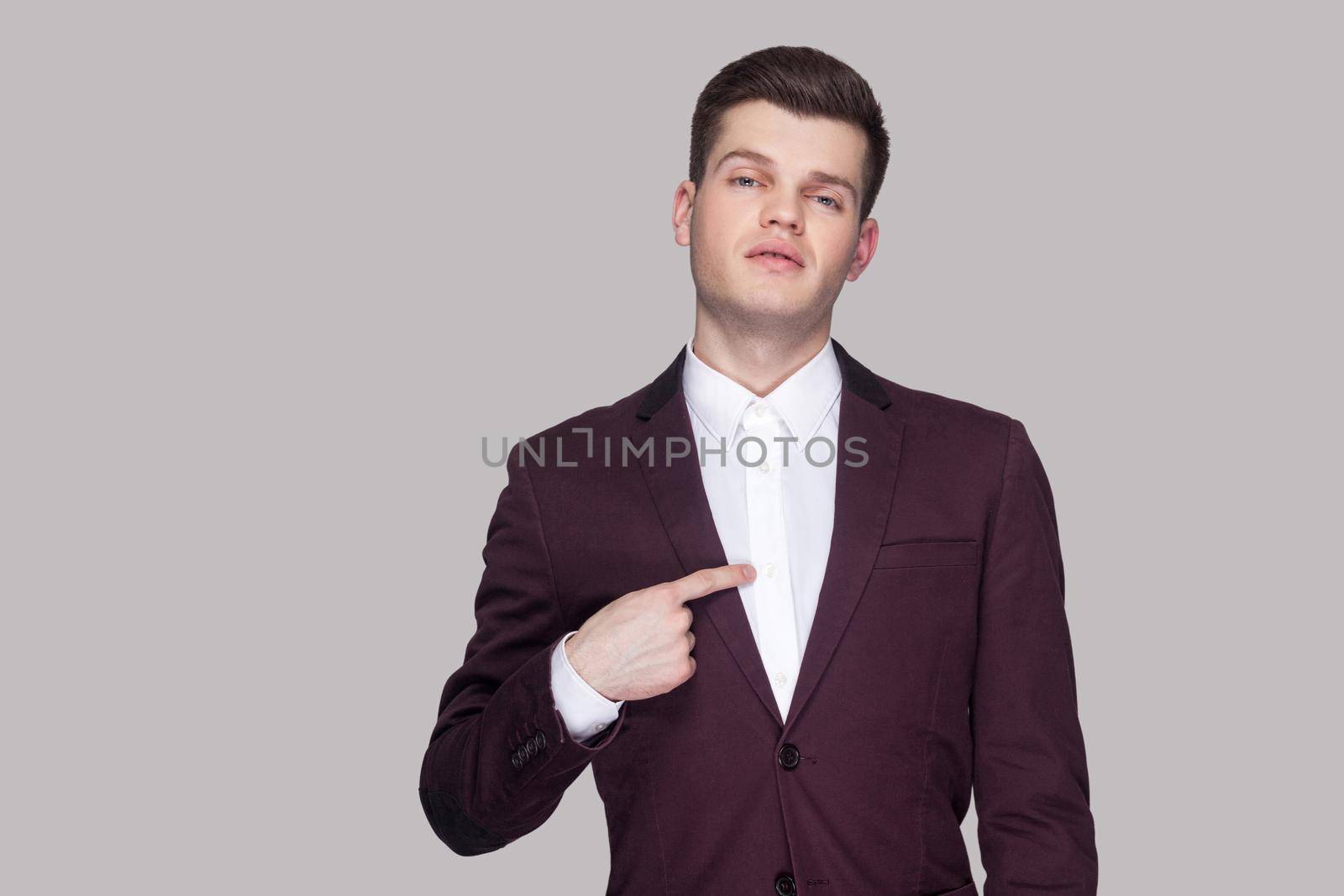 This is me. Portrait of proud handsome young businessman in violet suit and white shirt, standing, looking at camera and pointing himself and bragging. indoor studio shot, isolated on grey background