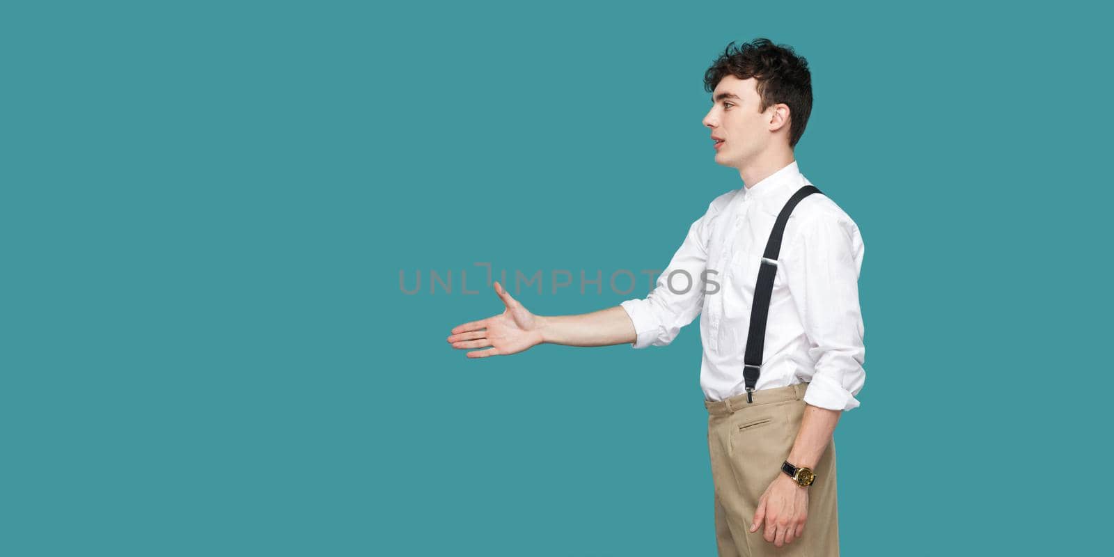 Profile side view portrait of happy handsome curly young businessman in classic casual white shirt standing and giving hand to greeting or handshake. indoor studio shot isolated on blue background.
