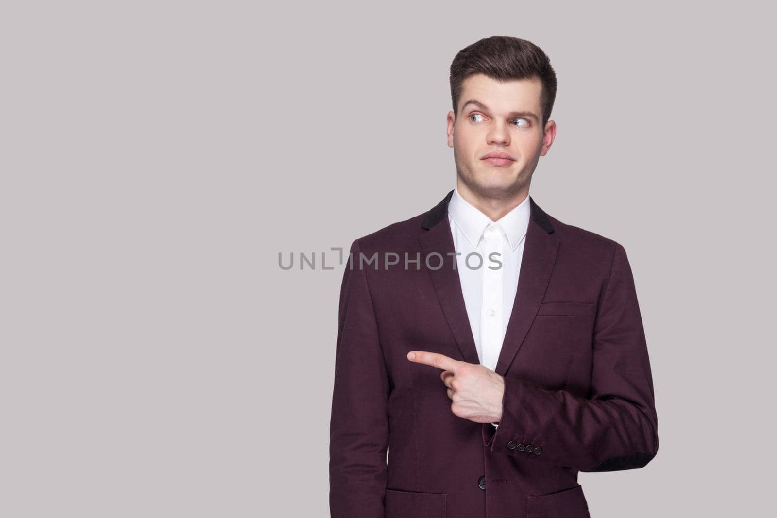 Portrait of funny handsome young man in violet suit and white shirt, standing, pointing and looking away with funny face and smiling. indoor studio shot, isolated on grey background.