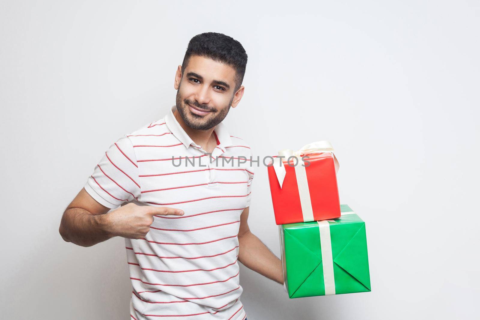 Portrait of cheerful attractive bearded young adult man in white t-shirt standing and holding two present box and pointing finger. Indoor, isolated, studio shot, white background
