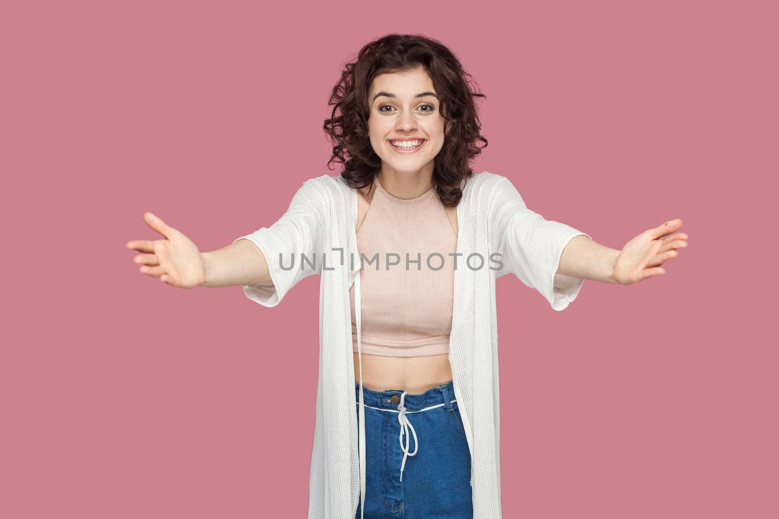 Portrait of happy beautiful brunette young woman with curly hairs in casual style standing with raised arms, want to hug, toothy smile and looking at camera. studio shot isolated on pink background.