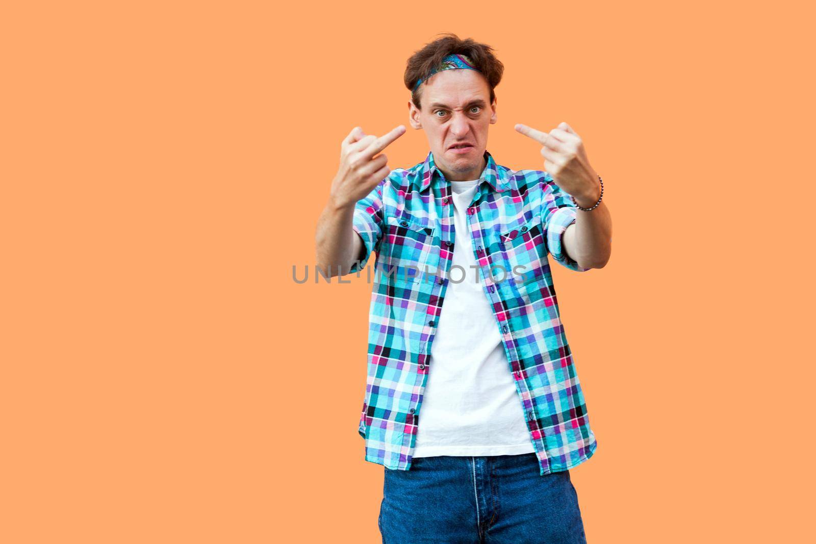 Portrait of angry young man in casual blue checkered shirt and headband standing with middle finger fuck sign looking at camera with aggressive face. indoor studio shot, isolated on orange background.