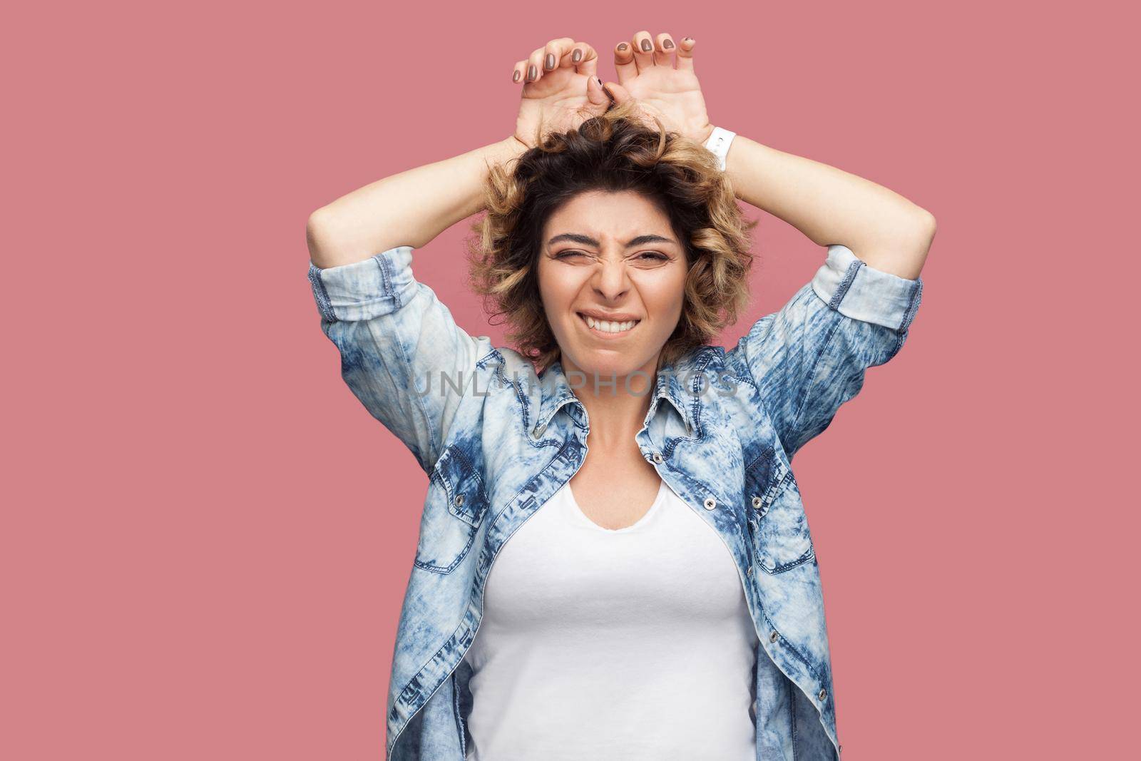 Portrait of funny crazy young woman with curly hair in casual shirt standing with funny face and bunny ears gesture hands on head and looking at camera. indoor studio shot, isolated on pink background