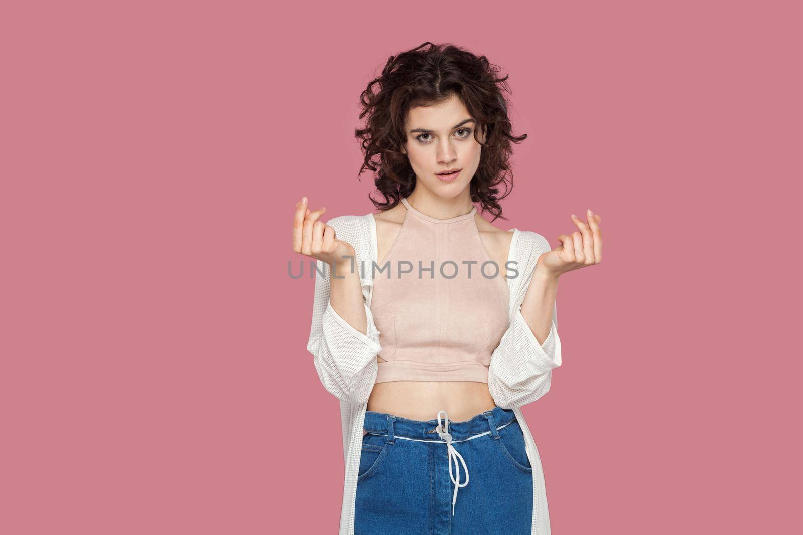 Portrait of beautiful brunette young woman with curly hairstyle in casual style standing, looking at camera and showing money or italian hand gesture. indoor studio shot isolated on pink background.