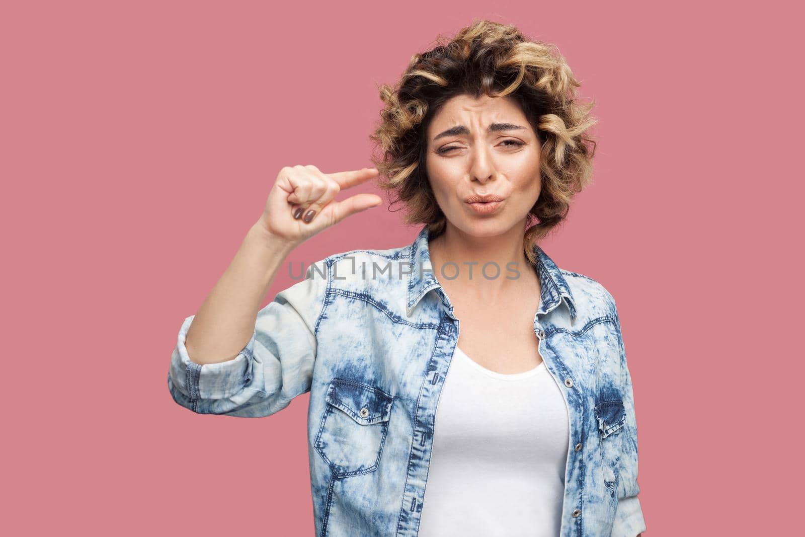 Give me a few more. Portrait of pleased worry young woman with curly hairstyle in casual blue shirt standing and showing a few gesture and pleading. indoor studio shot, isolated on pink background.