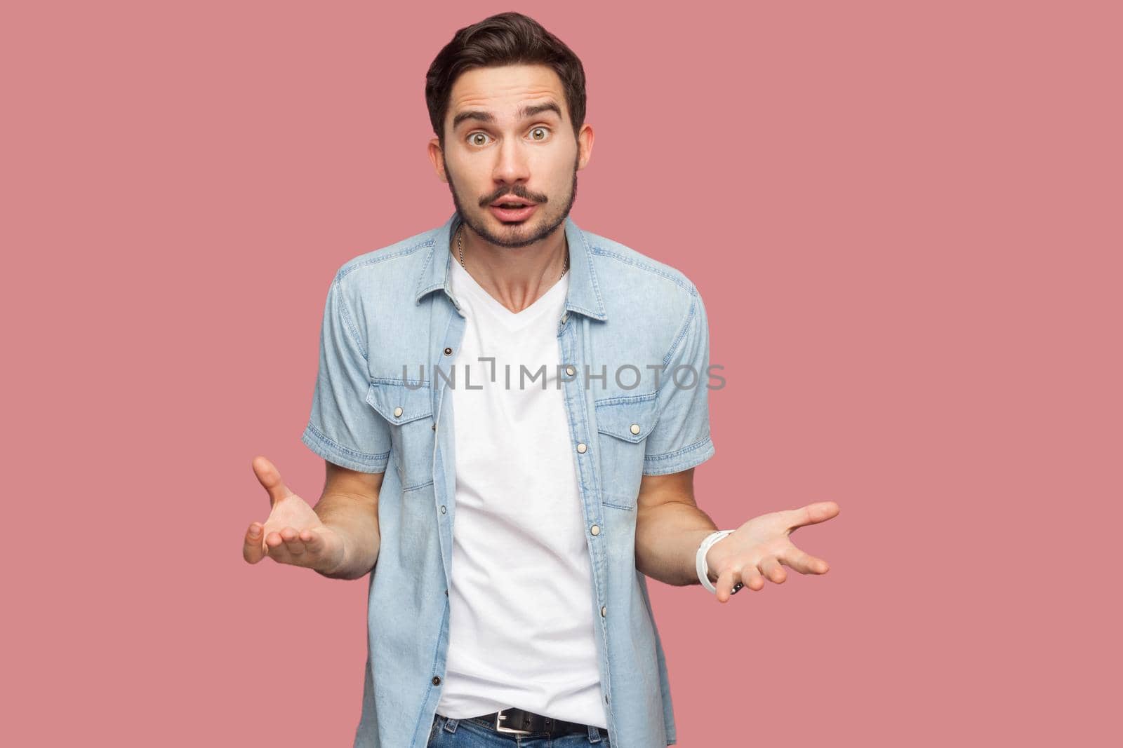 Portrait of angry shocked handsome bearded young man in blue casual style shirt standing and looking at camera and asking. indoor studio shot, isolated on pink background.