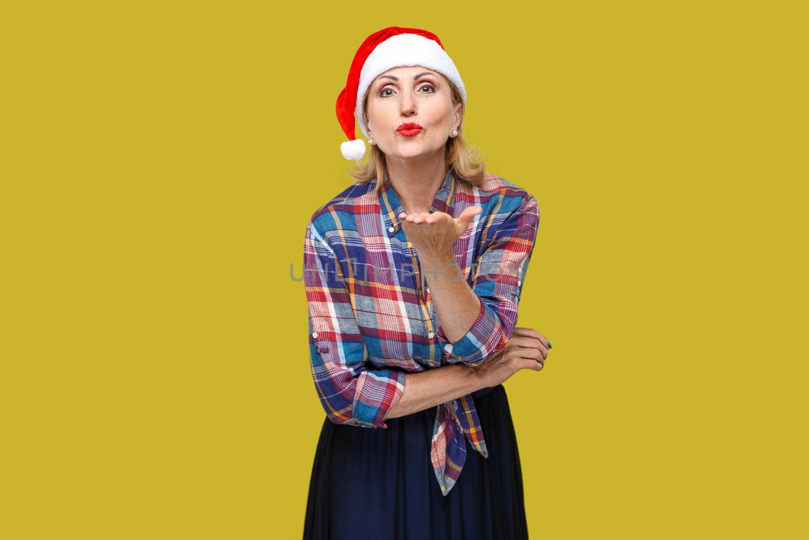 Kissing you! Portrait of kindly beautiful adult woman in red santa cap and checkered shirt standing and sending you air kiss, looking at camera. Indoor, studio shot, yellow background
