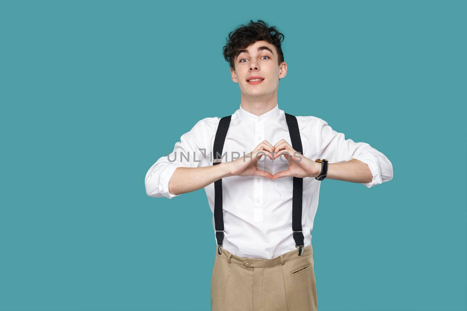 happy toothy smile man with heart hands sign gesture looking at camera. Portrait of handsome hipster curly young businessman in classic casual white shirt. studio shot isolated on blue background.