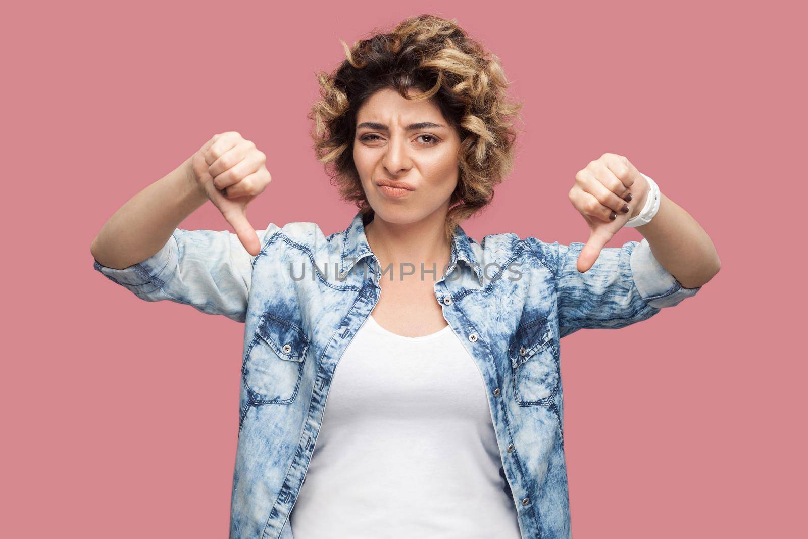 Dislike. Portrait of sad young woman with curly hairstyle in casual blue shirt standing, thumbs down and looking at camera with dissatisfied face. indoor studio shot, isolated on pink background.