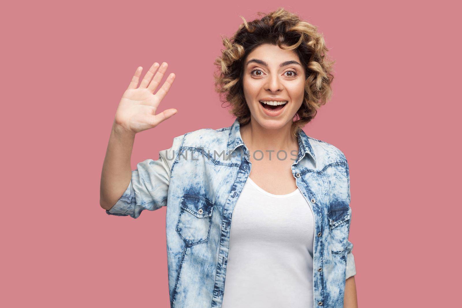 Hi. Portrait of surprised happy young woman with curly hairstyle in casual blue shirt standing, greeting, waving her hand and looking with toothy smile. indoor studio shot, isolated on pink background