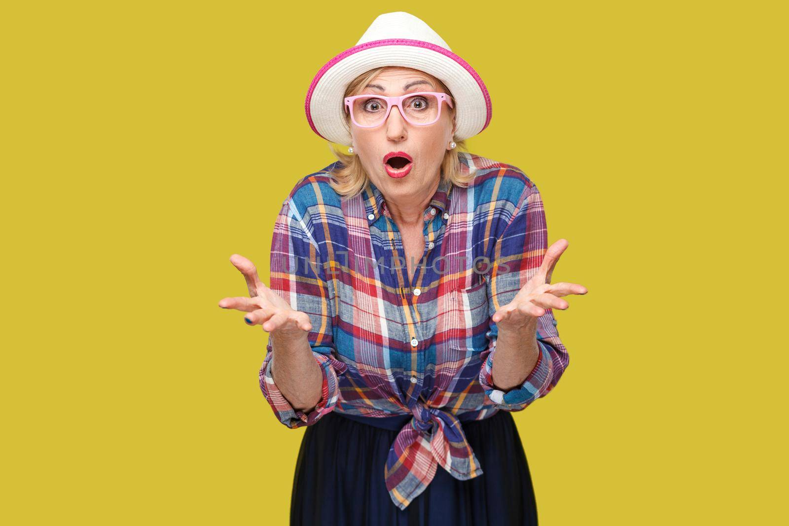 Portrait of surprised modern stylish mature woman in casual style with hat and eyeglasses standing, raised arms, looking at camera with shocked face. indoor studio shot isolated on yellow background.
