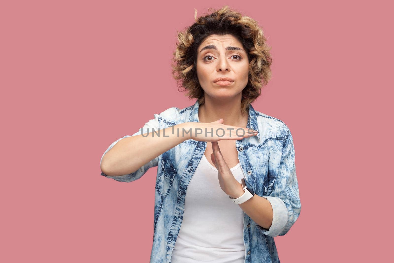 Timeout. I need more time. Portrait of worry pleased young woman with curly hairstyle in casual blue shirt standing with T gesture sign and looking at camera. studio shot, isolated on pink background
