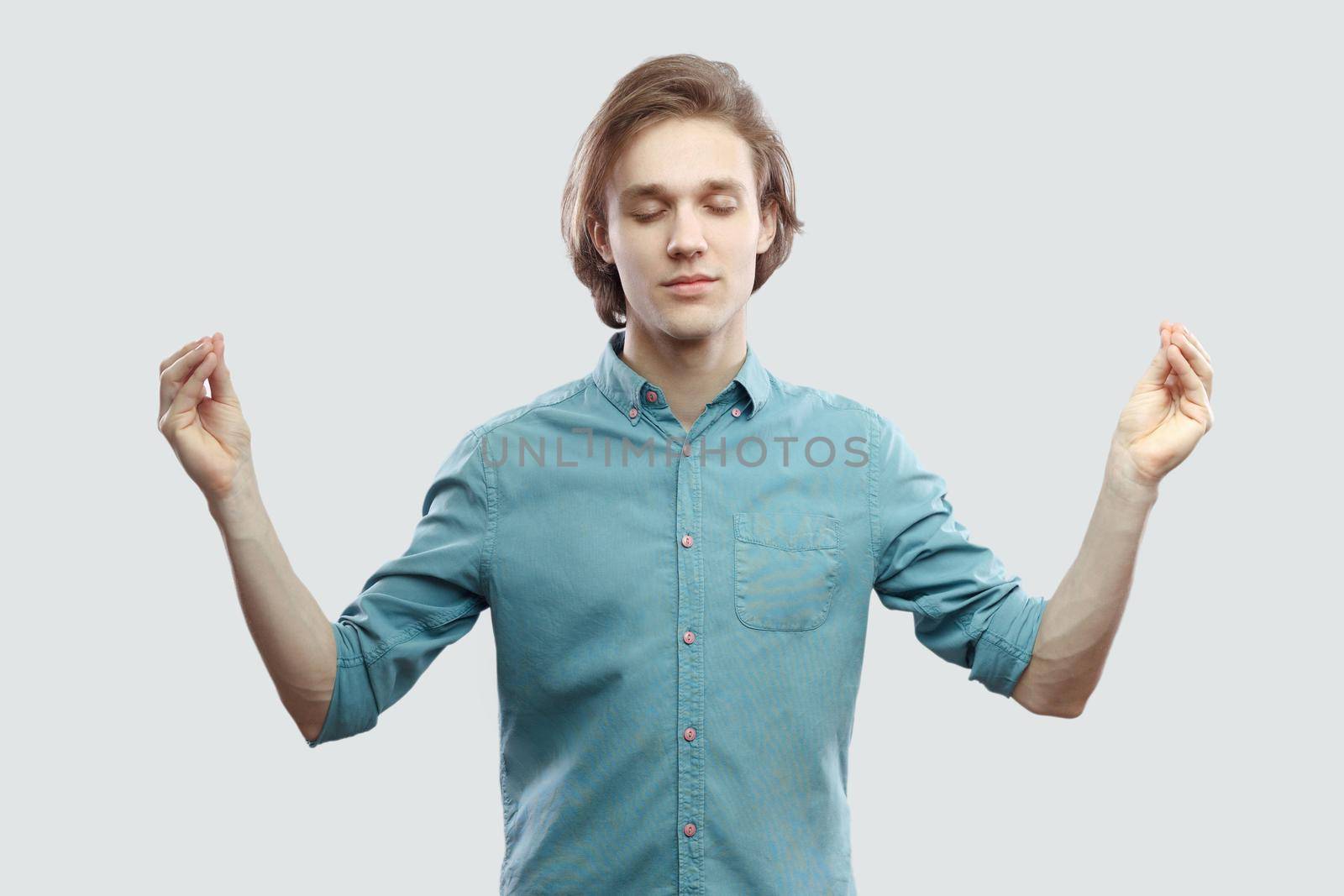 Portrait of calm handsome long haired blonde young man in blue casual shirt standing in yoga pose and meditating, and try to relax. indoor studio shot, isolated on light grey background.