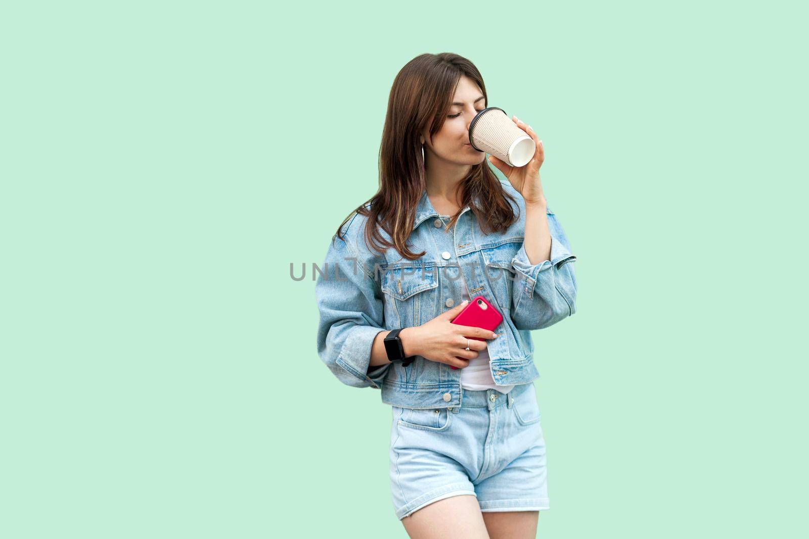 Portrait of beautiful young brunette woman in casual denim style standing and holding disposable cup, drinking and resting and enjoying. indoor studio shot, isolated on green background.