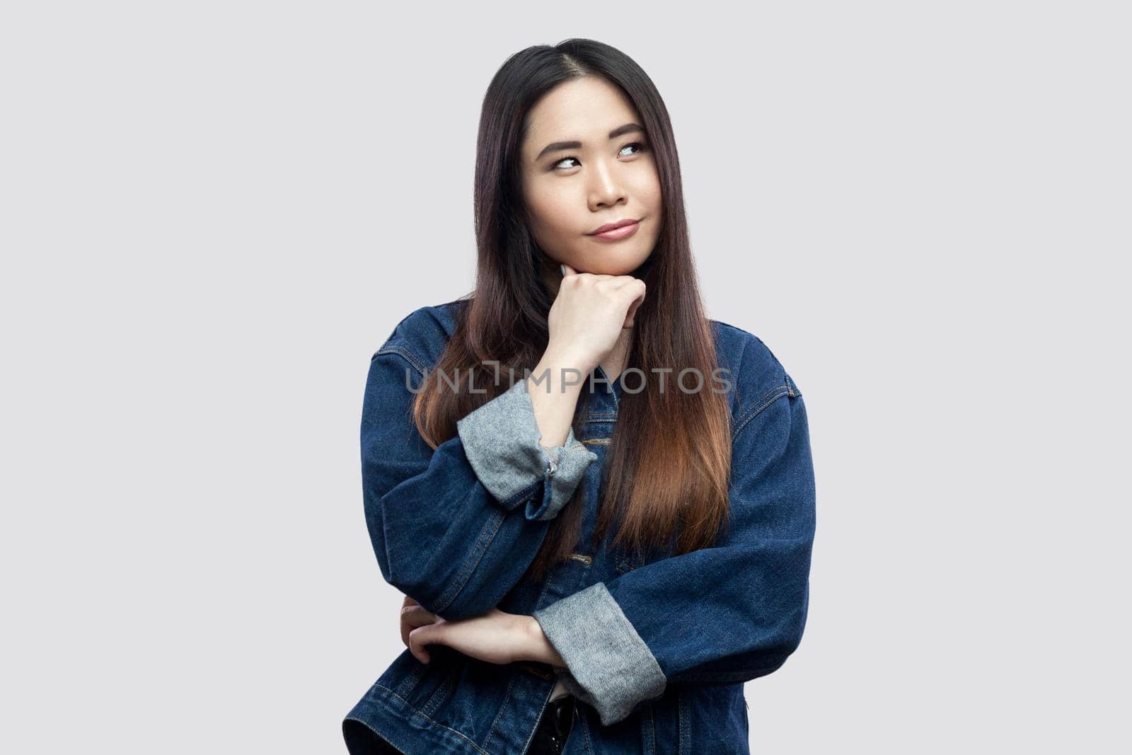 Portrait of thoughtful beautiful brunette asian young woman in casual blue denim jacket with makeup standing, confused looking away and thinking. indoor studio shot, isolated on light grey background.
