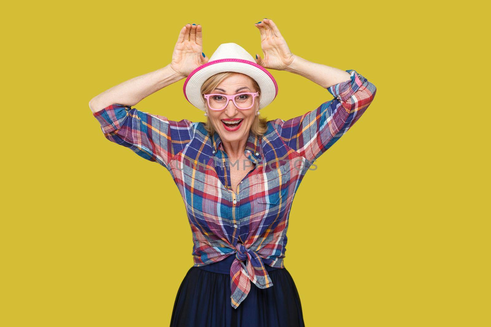 Portrait of funny modern stylish mature woman in casual style with hat and eyeglasses standing with bunny ears gesture and looking with toothy smile. indoor studio shot isolated on yellow background.