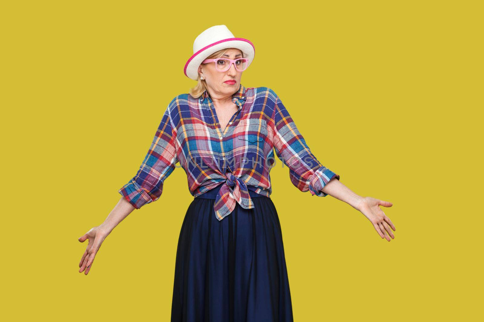 I don't know. Portrait of confused modern stylish mature woman in casual style with hat and eyeglasses standing, thinking, raised arms, looking away. indoor studio shot isolated on yellow background.