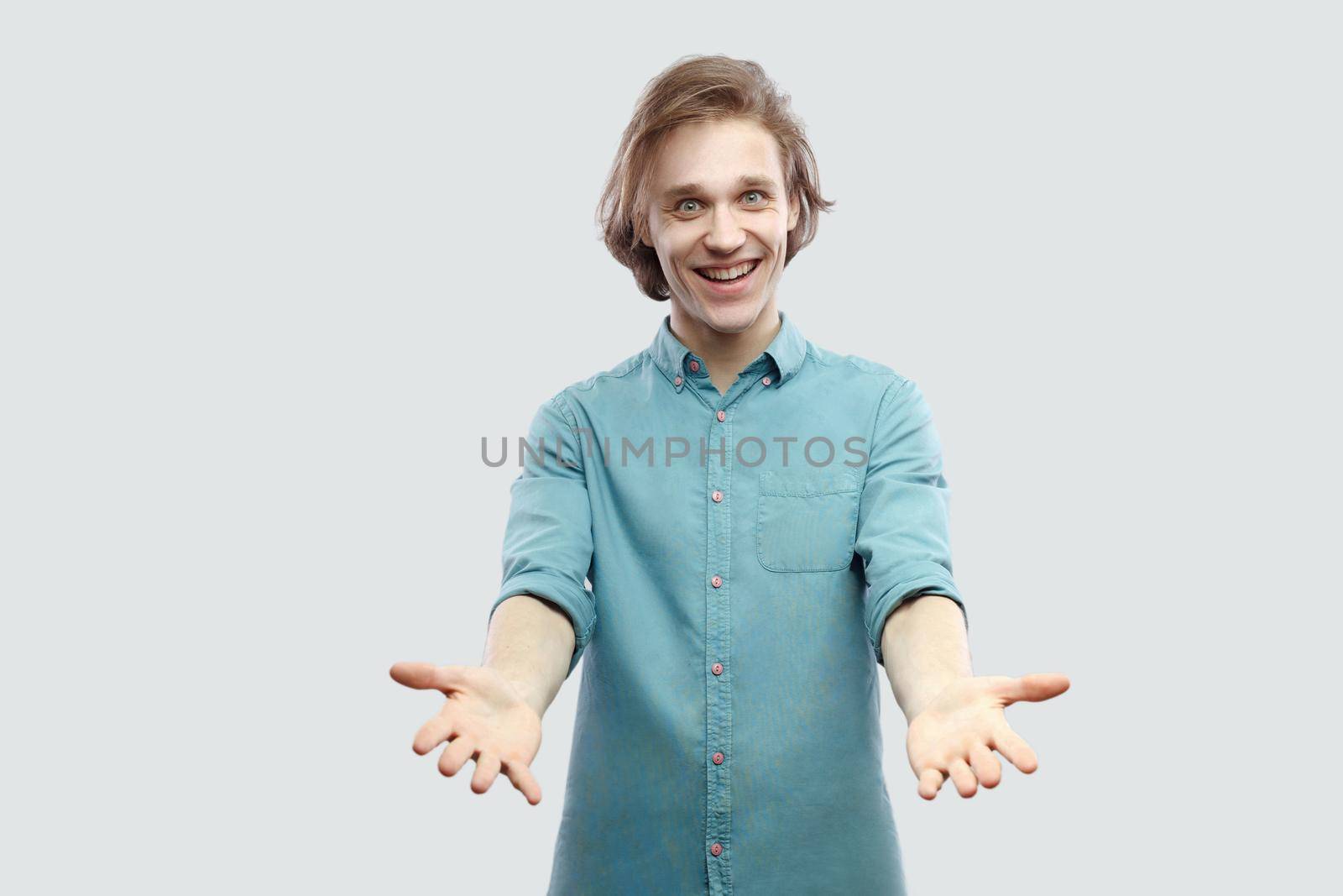 Portrait of happy genial handsome long haired blonde young man in blue casual shirt standing with raised arms and toothy smile looking at camera. indoor studio shot, isolated on light grey background.