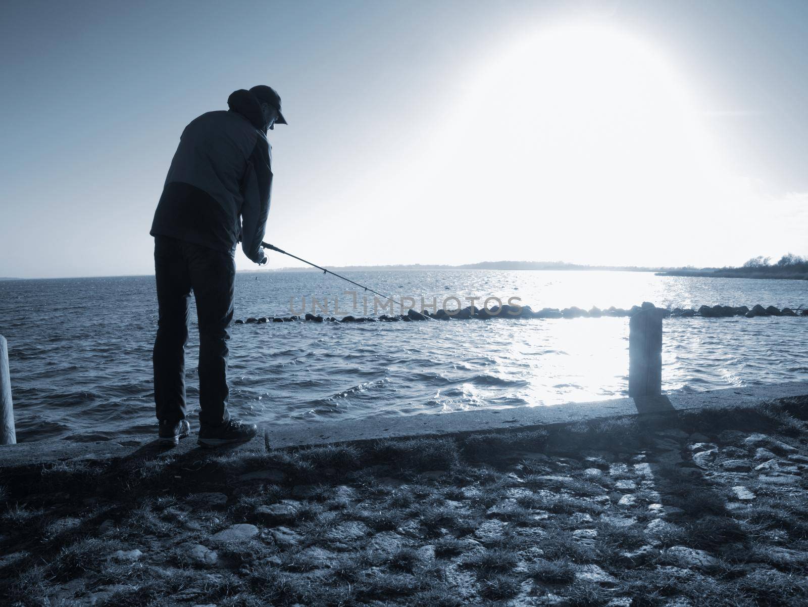 Fisherman with hat on the sea mole  with fish rod. Man standing above sea water and  fishing at sunset 